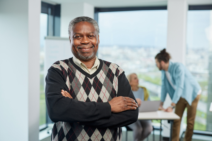 A smiling multicultural senior student is standing with arms crossed in a classroom and looking at the camera. Seniors with bachelor's degree concept.
